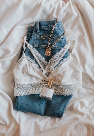 A pair of blue denim jeans is laid out on a white fabric. On top of the jeans, there is a white lace bralette. A gold chain necklace with a pendant and a small bottle of perfume are also placed on the jeans.