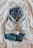 A pair of blue denim jeans is laid out on a white fabric. On top of the jeans, there is a white lace bralette. A gold chain necklace with a pendant and a small bottle of perfume are also placed on the jeans.