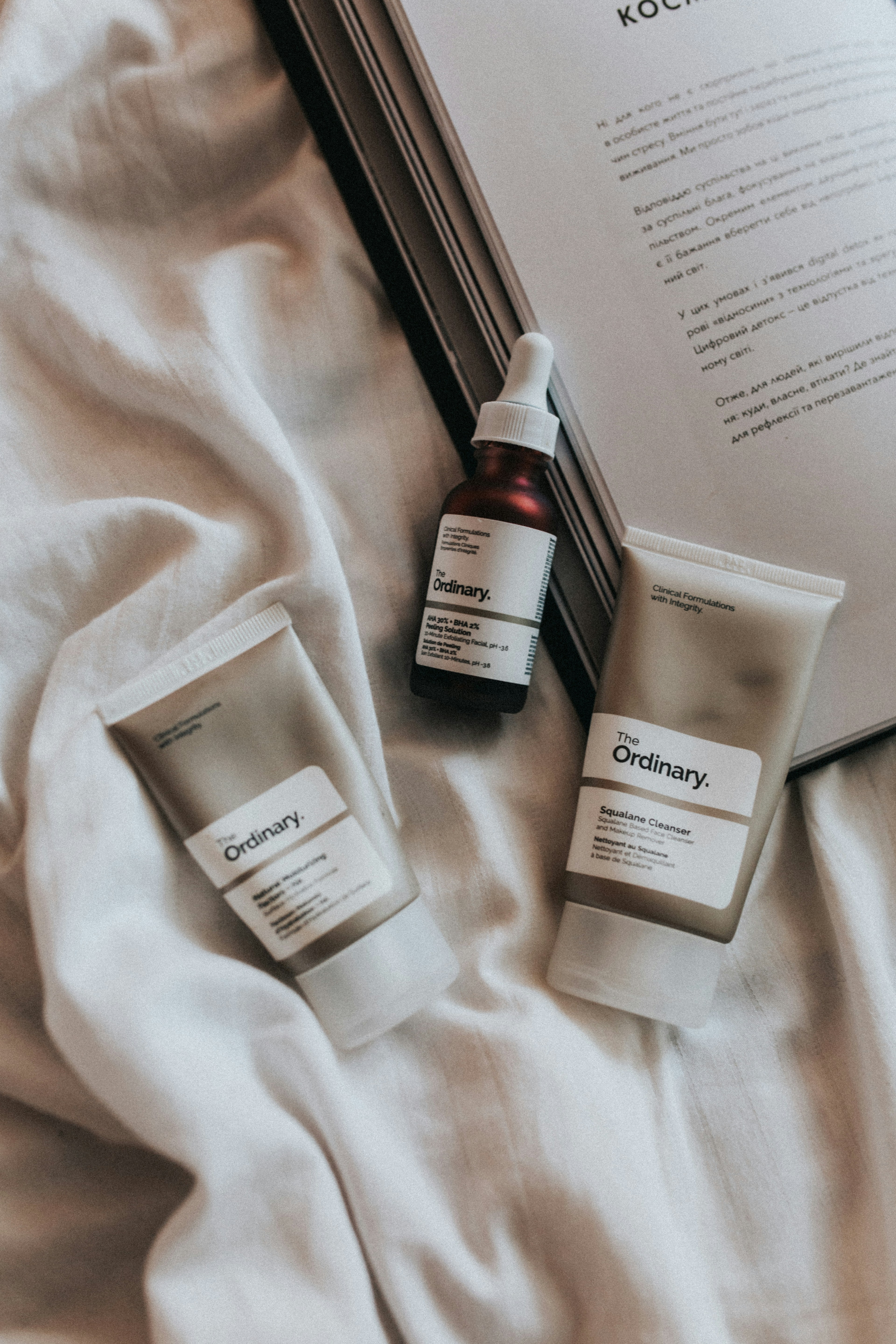 Flat lay of The Ordinary skincare products and an open book on white fabric.