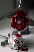 Two paper flowers, one red and one white with red edges, are placed on a surface. The red flower is positioned upright in an empty bottle with its intricate petals fully open. The white flower lies on the surface, surrounded by red liquid resembling spilled paint or ink. The backdrop is blurred, providing a focus on the detailed texture of the flowers.