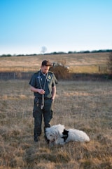 A person dressed in a dark uniform stands in a grassy field holding a leash. A large, fluffy white dog wearing a 'security' vest is sitting on the ground next to the person. The background includes a grassy hill with a lone tree and a clear blue sky.
