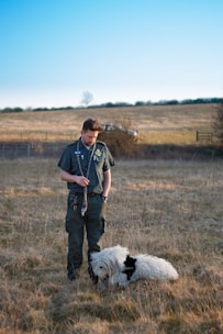 A person dressed in a dark uniform stands in a grassy field holding a leash. A large, fluffy white dog wearing a 'security' vest is sitting on the ground next to the person. The background includes a grassy hill with a lone tree and a clear blue sky.