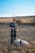 A person dressed in a dark uniform stands in a grassy field holding a leash. A large, fluffy white dog wearing a 'security' vest is sitting on the ground next to the person. The background includes a grassy hill with a lone tree and a clear blue sky.