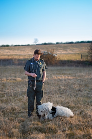 A person dressed in a dark uniform stands in a grassy field holding a leash. A large, fluffy white dog wearing a 'security' vest is sitting on the ground next to the person. The background includes a grassy hill with a lone tree and a clear blue sky.