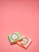 Two small wrapped chocolates lie on a pink surface. The wrapping includes elegant branding with the text 'Laduree Paris' and decorative golden embellishments. One wrapper is pastel green, and the other is a light pink.