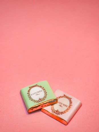 Two small wrapped chocolates lie on a pink surface. The wrapping includes elegant branding with the text 'Laduree Paris' and decorative golden embellishments. One wrapper is pastel green, and the other is a light pink.