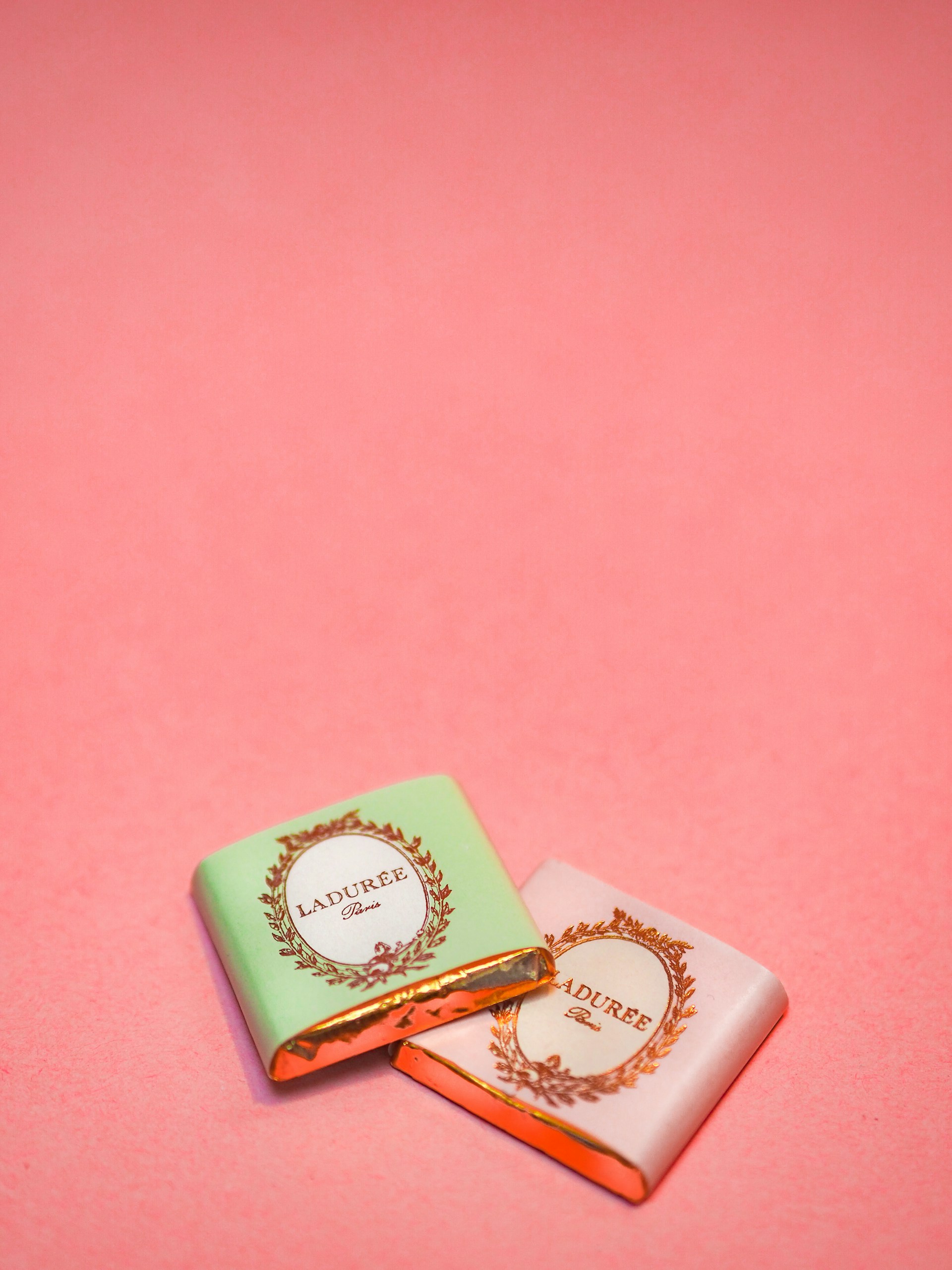 Two small wrapped chocolates lie on a pink surface. The wrapping includes elegant branding with the text 'Laduree Paris' and decorative golden embellishments. One wrapper is pastel green, and the other is a light pink.
