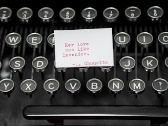 A vintage typewriter with a small white card placed on its keys. The card contains a typed message in red ink: 'Her love was like lavender. - L. Chouette'. The typewriter keys are metallic and circular with black centers and white letters.