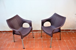 Two brown, woven-style chairs with metal legs are positioned on a terracotta-tiled floor against a plain white wall. The seating features a rounded square backrest and armrests with oval-shaped holes, giving them a modern and minimalist appearance.