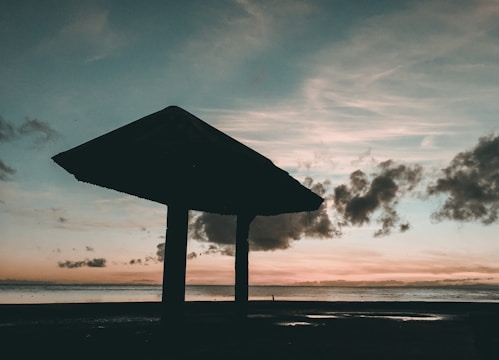 A silhouette of a shelter by the seaside against a colorful sunset sky. The horizon shows a blend of orange and pink hues while darker clouds drift across, creating a picturesque scene. The foreground features a darkened structure with two visible supporting columns.