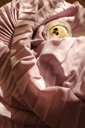 Soft, flowing pink satin fabric is draped elegantly, creating graceful folds and shadows. A small glass jar containing a creamy substance topped with coffee beans is placed on the fabric, adding a touch of luxury and indulgence.