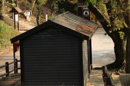 A small wooden shed with a sloped roof is situated beside a dirt pathway. Fallen leaves can be seen on the roof, indicating a natural, rustic setting. There are trees nearby, and a pathway leads up with steps on the left side. Signs are visible in the background, suggesting a park or nature trail area.