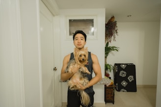 A person is standing indoors, holding a small dog that appears to be a Yorkshire Terrier. The background is a simple interior with white walls, a door, and a small table with a decorative plant. A piece of folded black fabric with white patterns is leaning against the wall.
