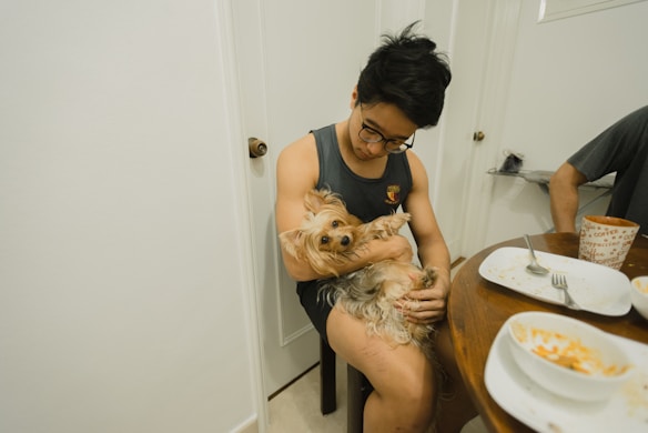 A person with glasses is sitting and holding a small, fluffy dog in their lap. The setting appears to be a dining area with a table that has empty plates, a bowl, and utensils. The person is wearing a sleeveless shirt and has a relaxed demeanor.