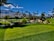 A lush green landscape featuring a well-manicured golf course with vibrant grass and a variety of plants. Tall palm trees are scattered throughout the area, with mountains visible in the background under a clear blue sky. In the foreground, a small pond is surrounded by flowers and shrubs.