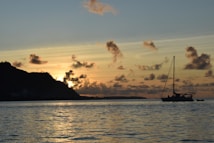 A peaceful seascape during sunset with a sailboat anchored in the water. The sky features scattered clouds with shades of yellow, orange, and hints of purple. In the background, there is a silhouette of a hilly landscape. The water reflects the colors of the sky, enhancing the serene atmosphere.