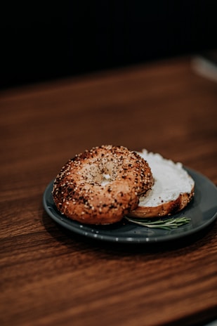 A bagel sliced in half, topped with a thick layer of cream cheese, is placed on a dark blue plate. The bagel is sprinkled with a variety of seeds, including sesame and poppy seeds. The plate rests on a wooden surface, and a small sprig of rosemary is visible beside the bagel on the plate.