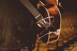 A close-up view of a person holding an acoustic guitar with a patterned strap. The guitar features a dark wood finish. The person appears to be on a stage, possibly preparing for a performance, as indicated by the audio equipment and cables connected to the guitar.