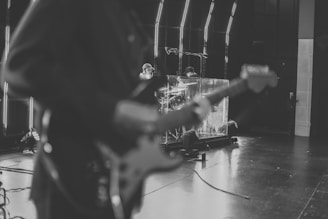 A monochrome image of a band setup with a guitar player in the foreground and a drummer behind a transparent sound barrier on a stage, surrounded by equipment and stage lights.