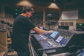 A person is adjusting controls on a large audio mixing console inside a spacious room, possibly a theater or auditorium. The environment is dimly lit, and the background has numerous chairs and some overhead lighting equipment. There is a large screen on the wall, and other technical equipment is visible.