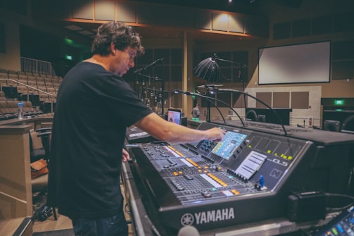 A person is adjusting controls on a large audio mixing console inside a spacious room, possibly a theater or auditorium. The environment is dimly lit, and the background has numerous chairs and some overhead lighting equipment. There is a large screen on the wall, and other technical equipment is visible.