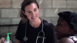 A healthcare professional is attentively interacting with a person. The professional is wearing a black shirt with a stethoscope around the neck, suggesting a medical or caregiving setting. A bottle of hand sanitizer is visible on the table.