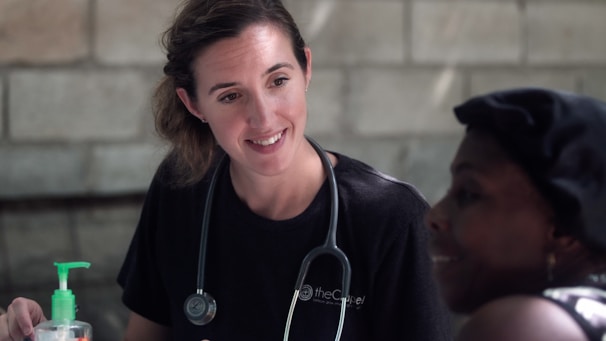 A healthcare professional is attentively interacting with a person. The professional is wearing a black shirt with a stethoscope around the neck, suggesting a medical or caregiving setting. A bottle of hand sanitizer is visible on the table.