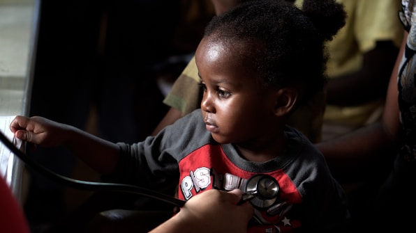 A young child is seated, looking intently to the side. They are wearing a red and gray shirt with a graphic print. A person is holding a stethoscope to the child's chest, suggesting a medical examination. The background is dimly lit, focusing the light on the child's face and creating a contrast with the darker surroundings.