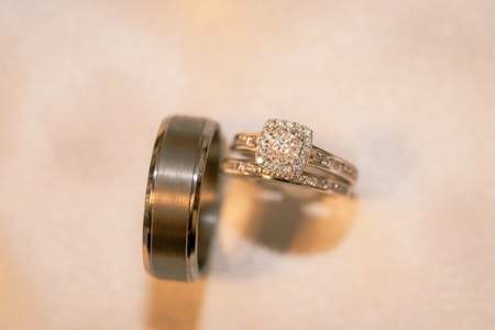 A pair of gold rings with intricate detailing. One is a wide band with a smooth finish, while the other is a more delicate ring featuring a square-shaped diamond surrounded by smaller diamonds, likely an engagement ring.