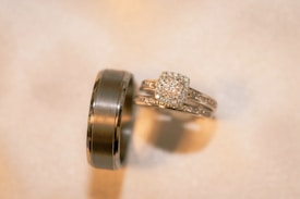 A pair of gold rings with intricate detailing. One is a wide band with a smooth finish, while the other is a more delicate ring featuring a square-shaped diamond surrounded by smaller diamonds, likely an engagement ring.