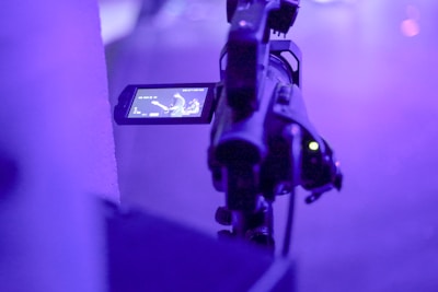 A video camera is set up to film a performance. The camera screen is visible, showing a musician playing a guitar onstage. The scene is illuminated with purple lighting, creating a moody and atmospheric ambiance.