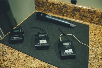 Several Sennheiser wireless microphone transmitters and receivers are laid out on a black mat on a countertop. There is also a handheld microphone positioned next to them. The devices appear to have display screens and numbered labels.