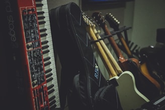 A collection of musical instruments including a red and white keyboard and several guitars with different body colors, leaning against a wall. There are also guitar cases in the foreground, creating a cozy studio or rehearsal room atmosphere.