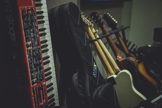 A collection of musical instruments including a red and white keyboard and several guitars with different body colors, leaning against a wall. There are also guitar cases in the foreground, creating a cozy studio or rehearsal room atmosphere.