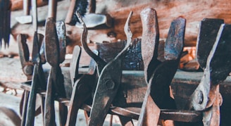 A collection of old, rusted metal tools, likely used for blacksmithing or metalworking, are densely arranged against a wooden backdrop. The tools exhibit notable wear and patina, indicative of long-term use.