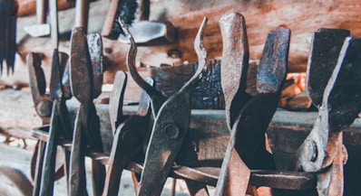 A collection of old, rusted metal tools, likely used for blacksmithing or metalworking, are densely arranged against a wooden backdrop. The tools exhibit notable wear and patina, indicative of long-term use.