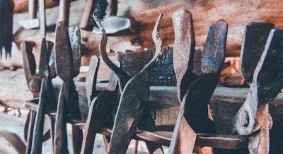 A collection of old, rusted metal tools, likely used for blacksmithing or metalworking, are densely arranged against a wooden backdrop. The tools exhibit notable wear and patina, indicative of long-term use.