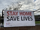 A large banner is displayed outdoors on a black metal fence, with the words 'STAY HOME SAVE LIVES' in bold red and black lettering. The lower part of the banner includes the logo for Hammersmith & Fulham Council and a URL for coronavirus information. The banner is set against a backdrop of a cloudy sky and some trees.