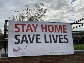 A large banner is displayed outdoors on a black metal fence, with the words 'STAY HOME SAVE LIVES' in bold red and black lettering. The lower part of the banner includes the logo for Hammersmith & Fulham Council and a URL for coronavirus information. The banner is set against a backdrop of a cloudy sky and some trees.