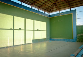 A large indoor court with high walls painted green and lined with yellow, situated under a metallic roof that allows natural light to create shadows on the walls. The court has markings on the floor and the walls reflect a shadow pattern creating a grid-like effect.