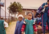 A group of young children wearing uniforms stand outdoors near a brick building and chain-link fence. Some children are playing, while adults and older children are wearing casual clothing. A tree and various toys are visible in the background.