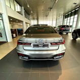 A silver luxury sedan is positioned in a modern car showroom with large glass windows allowing natural light to fill the space. The vehicle is facing away from the viewer, showcasing its sleek design and detailed rear end, including distinct taillights and the model badge '740Li'. Other vehicles and office areas are visible in the background.