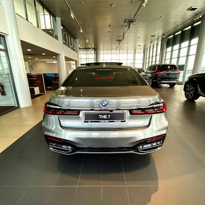 A silver luxury sedan is positioned in a modern car showroom with large glass windows allowing natural light to fill the space. The vehicle is facing away from the viewer, showcasing its sleek design and detailed rear end, including distinct taillights and the model badge '740Li'. Other vehicles and office areas are visible in the background.