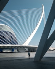 A futuristic architectural structure features sweeping geometric lines and a prominent arch with cables that resemble a harp. A lone person stands on the paved area beneath the structure, adding a sense of scale. The sky is clear and blue, creating a stark contrast with the white and blue tones of the building.