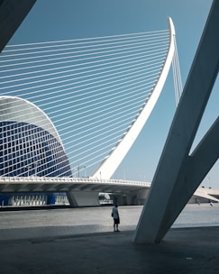 A futuristic architectural structure features sweeping geometric lines and a prominent arch with cables that resemble a harp. A lone person stands on the paved area beneath the structure, adding a sense of scale. The sky is clear and blue, creating a stark contrast with the white and blue tones of the building.