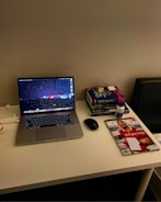 A neatly organized desk featuring a laptop with a colorful screensaver, a mouse beside it, and books stacked to the right. On top of the books is a pair of wireless earbuds and a bottle of vitamins. A magazine with a cover displaying a person in a red shirt lies adjacent to the stack of books. The setup conveys a tidy and functional workspace.