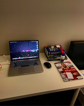A neatly organized desk featuring a laptop with a colorful screensaver, a mouse beside it, and books stacked to the right. On top of the books is a pair of wireless earbuds and a bottle of vitamins. A magazine with a cover displaying a person in a red shirt lies adjacent to the stack of books. The setup conveys a tidy and functional workspace.