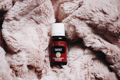 A small bottle of essential oil is placed on top of a fluffy, beige blanket. The bottle is dark with a white cap and a label indicating it is from Young Living. The text on the label reads 'Thieves' and includes other details about the product.