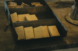 A collection of small, vintage-style booklets with faded covers arranged neatly within a metal tray resting on a rustic wooden table. Each booklet features minimal printed text, giving a nostalgic and handcrafted feel.