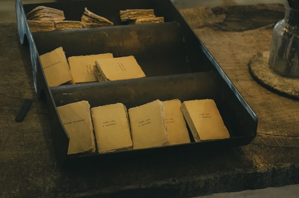 A collection of small, vintage-style booklets with faded covers arranged neatly within a metal tray resting on a rustic wooden table. Each booklet features minimal printed text, giving a nostalgic and handcrafted feel.